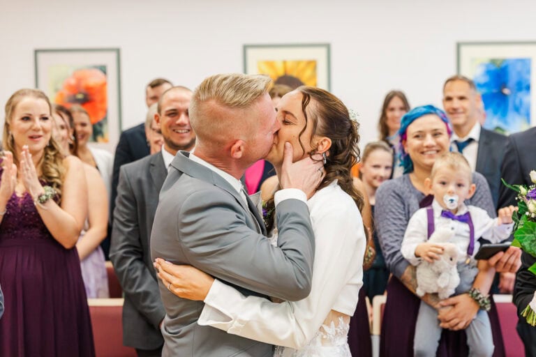 Hochzeitspaar beim Kuss, applaudierende Gäste im Hintergrund.